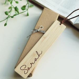 May include: A personalized wooden bookmark with the name "Sarah" engraved in brown lettering. The bookmark is tied with a silver ribbon and comes in a brown cardboard box.