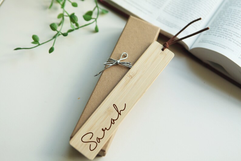 May include: A personalized wooden bookmark with the name "Sarah" engraved in brown lettering. The bookmark is tied with a silver ribbon and comes in a brown cardboard box.