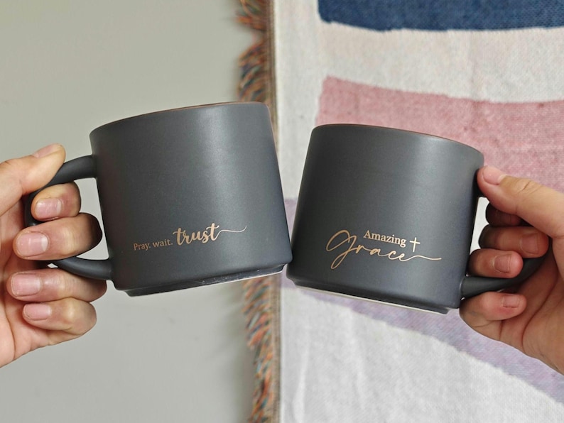 May include: Two matte charcoal gray ceramic mugs with gold lettering. One mug features the text "Pray, wait. trust" and the other features the text "Amazing grace" with a small cross symbol. Both mugs have a smooth finish and a handle.