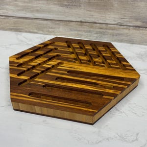 May include: A hexagonal wooden trivet with a geometric design. The trivet is made of alternating strips of light and dark wood, creating a striped pattern. The edges are a lighter colour, and the trivet has a raised border.