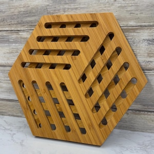 May include: A hexagonal bamboo trivet with a natural wood finish. The trivet has a series of rectangular and diagonal cutouts, creating an open, geometric design. This kitchen accessory is designed to protect surfaces from hot cookware.