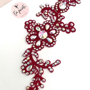 May include: A burgundy-coloured, floral-shaped applique with iridescent rhinestones. The applique is made of a crocheted material and features a central flower design with surrounding smaller rhinestones. The design is from "En pointe designs".
