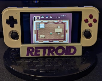 May include: A beige handheld gaming console with a black and white screen displaying a pixelated video game. The console has a purple stand with the word "RETROID" printed on it. The console is sitting on a black surface.