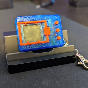 May include: A blue and orange digital pet keychain with a screen displaying a pixelated image. The keychain is attached to a silver metal clasp.