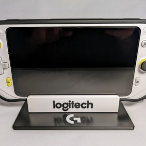 Puede incluir: Consola de juegos portátil blanca y negra con pantalla negra, detalles amarillos y soporte negro con el logotipo de Logitech. La consola tiene botones y joysticks. El soporte es negro.