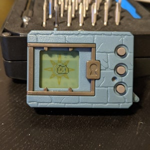 Puede incluir: Un juguete Digivice azul claro con una pequeña pantalla que muestra una imagen pixelada. El juguete tiene forma rectangular con un diseño texturizado en forma de ladrillo. Cuenta con tres botones grises y un detalle de ojo de cerradura.