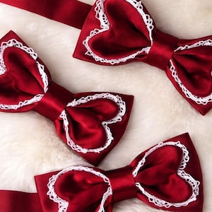 Peut inclure: Trois nœuds papillon en satin rouge avec des centres en forme de cœur et une bordure en dentelle blanche. Les nœuds papillon sont disposés sur une surface douce et blanche. Les nœuds ont un design classique.
