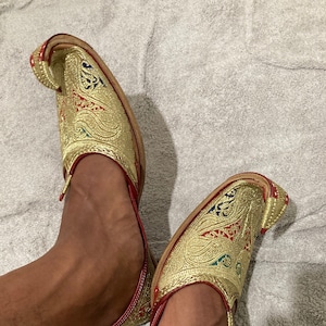 Puede incluir: Un par de zapatos bordados en oro con detalles rojos. Los zapatos tienen una punta puntiaguda y una parte superior curva. Los zapatos están puestos en los pies de una persona.