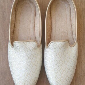 May include: A pair of white and gold embroidered flat shoes. The shoes have a diamond pattern and a pointed toe.