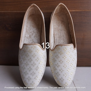 May include: A pair of white and gold patterned slippers with a brown trim. The size 13 is written on the side of the slippers.