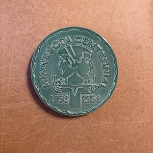 May include: A green ceramic plate with a white design commemorating the Minnesota Centennial. The design features two stylised faces, a cityscape, and the years 1858 and 1958.