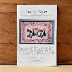 May include: A white instructional booklet titled Spring Fever featuring a quilted wall hanging with a central row of colorful tulips on a light background, surrounded by a floral border. Below, a black and white line drawing of tulips is shown.