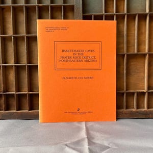 May include: An orange book titled "Basketmaker Caves in the Prayer Rock District, Northeastern Arizona" by Elizabeth Ann Morris. The book is part of the Anthropological Papers of the University of Arizona, Number 25. The book is on a gray surface.
