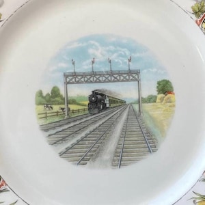 May include: White ceramic plate with a central illustration of a vintage steam train traveling on tracks under a bridge. The plate has a decorative floral and leaf border in yellow, green, and red.