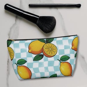 Può includere: Un beauty case con motivo limone, un grande pennello nero per il trucco e un piccolo pennello nero per il trucco. La borsa presenta uno sfondo a scacchi azzurro e bianco con illustrazioni di limoni e foglie verdi. La borsa ha una cerniera dorata.