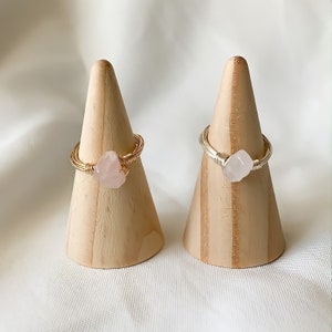 May include: Two gold wire wrapped rings with pink rose quartz stones. The rings are displayed on wooden ring cones.