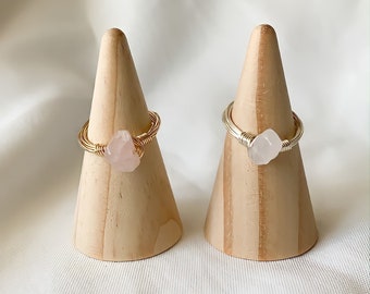 May include: Two gold wire wrapped rings with pink rose quartz stones. The rings are displayed on wooden ring cones.