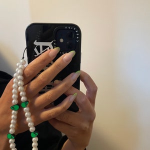 May include: A black phone case with a white design that says "CASEIFY" around the camera lens. A white pearl necklace with green alien face charms is attached to the phone case.