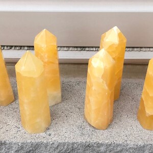 May include: A collection of 12 yellow calcite crystal points, each with a unique, faceted shape. The crystals are arranged in two rows on a light grey surface.