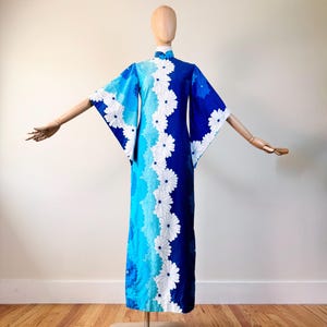 May include: A blue, white, and turquoise floral print dress with wide sleeves. The dress features a high collar and a vertical row of white daisy-like flowers down the front. The dress is displayed on a wooden mannequin.