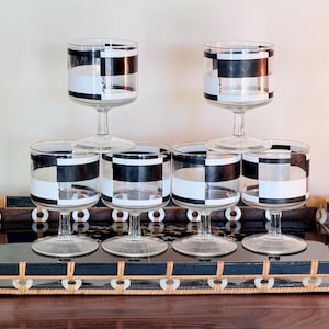 May include: A set of six clear glass goblets featuring a bold black and white checkered pattern around the bowl, arranged on a rectangular black tray with a woven rattan border and light-colored circular accents along the outer frame.