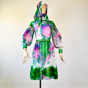 May include: A vintage floral dress with a matching headscarf on a mannequin. The dress features a green, white, pink, and purple floral pattern, long sleeves, and a green belt. The skirt is knee-length.