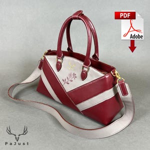 May include: A structured handbag in deep burgundy and light gray with two top handles and a detachable shoulder strap. The bag features a diagonal color-blocked design, a small floral graphic, and a gold logo. It includes a zipper closure.