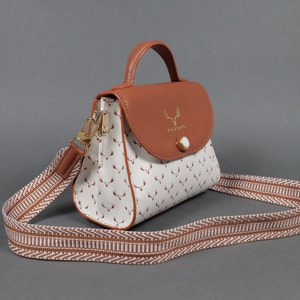 May include: A small brown and white handbag with a top handle and a long adjustable strap. The bag has a white background with a pattern of small white deer heads and a brown flap with the word "Palust" in gold lettering.