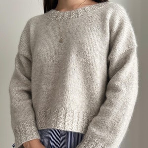 Op de afbeelding: Een lichtgrijze gebreide trui met een licht ingekorte zoom en lange mouwen. De trui heeft een delicaat kabelbreipatroon rond de hals en manchetten.