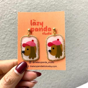 May include: Gold dangle earrings with a white background and a cartoon dog wearing a pink hat. The earrings are labeled "The Lazy Panda Studio" and include the social media handles @thelazypanda_studio and thelazypandastore.etsy.com.