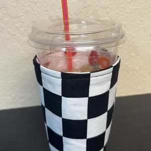 May include: A clear plastic cup filled with a pink beverage, topped with a clear lid and a pink and orange straw. The cup is wrapped in a black and white checkered fabric sleeve. The drink appears to contain fruit and gummy candies.