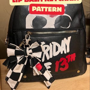 May include: Black backpack with a "Friday the 13th" design in white and red. A black and white checkered bow with a red lip balm keychain hangs from the zipper. The text "LIP BALM KEYCHAIN PATTERN" is at the top.