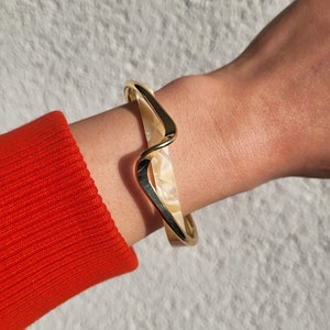 Könnte beinhalten: Goldfarbenes Armband mit Wellendesign und cremefarbenem, marmoriertem Inlay. Das Armband wird am Handgelenk getragen, mit einem roten Ärmel im Eck. Das Design des Armbands ist modern und elegant.