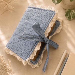 May include: A quilted fabric book cover in a light blue color with a small dark blue floral pattern. The cover features white eyelet lace trim along the edges and a matching fabric tie closure. The cover is wrapped around a thick book.