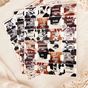 May include: A set of adhesive Bible index tabs featuring a cowhide print pattern in shades of brown, black, and white. Each tab is labeled with the name of a book of the Bible in black text, including Genesis, Exodus, Leviticus, Numbers, and others.