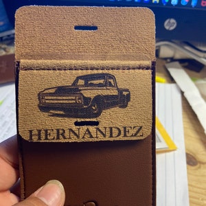 May include: A brown leather wallet with a flap that has a black and white image of a pickup truck and the text "HERNANDEZ" engraved on it. The wallet has a brown leather flap with a small hole in the top. The bottom of the wallet has the word "GATORISH" embossed on it.