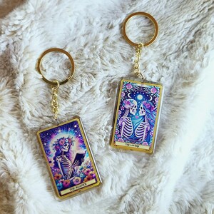 May include: Two rectangular keychains with gold-colored frames and key rings. Each keychain features a tarot card design with skeleton figures and floral accents. One reads "The Reader," the other "The Lovers."