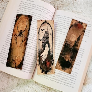 May include: Three gothic-themed bookmarks with illustrations of a spider, haunted house, and a moonlit landscape. The bookmarks are placed on an open book, creating a spooky aesthetic.