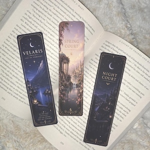 May include: Three rectangular bookmarks resting on an open book. The left bookmark features a dark blue night scene with the text Velaris City of Starlight. The center bookmark shows a light landscape with the text Spring Court. The right bookmark depicts a dark night scene with the text Night Court.