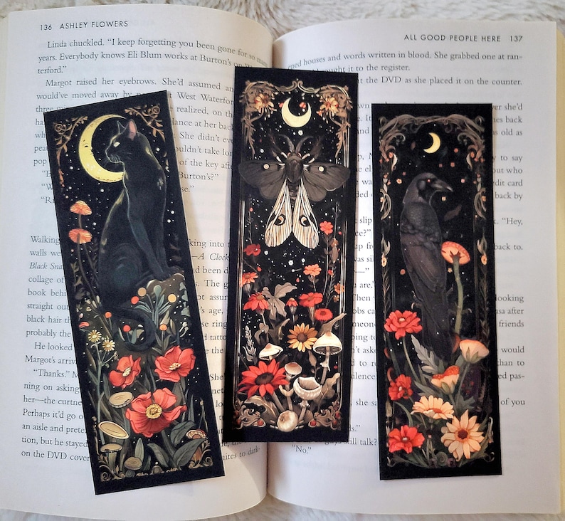 May include: Three black bookmarks with a dark background and a crescent moon in the top left corner. Each bookmark features a different creature: a black cat, a moth, and a raven. The creatures are surrounded by red and yellow flowers and mushrooms.