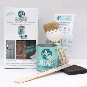 May include: A kit for creating weathered faux effects on furniture and other projects. The kit includes a container of Saltwash, a paintbrush, a measuring cup, a wooden stirring stick, and a sponge. The packaging features a woman holding a paintbrush and the text "Saltwash".