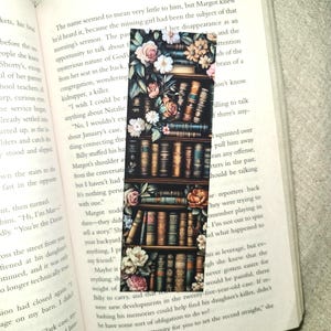 May include: A close-up of a book bookmark featuring a vintage illustration of a bookshelf filled with books and flowers. The bookmark is printed on thick paper and has a floral design with white, pink, and yellow flowers.