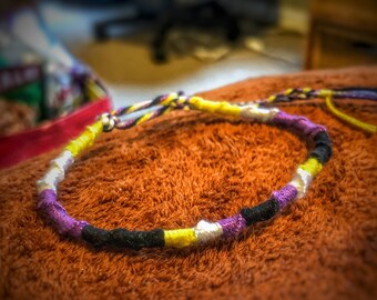 Könnte beinhalten: Ein lila, gelbes, schwarzes und weißes Freundschaftsarmband aus Schnur. Das Armband liegt auf einer braunen, flauschigen Oberfläche.
