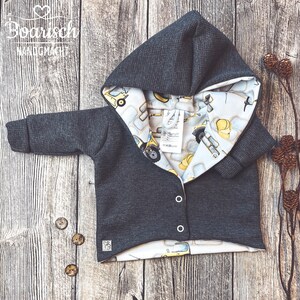 Könnte beinhalten: Eine dunkelgraue Kapuzenjacke mit einem weißen und gelben Baumaschinen-Druckfutter. Die Jacke hat einen Knopfverschluss und eine Kapuze.
