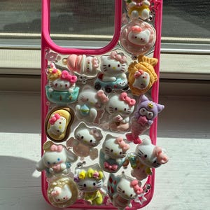 May include: A pink phone case adorned with multiple 3D Hello Kitty figurines in diverse poses and outfits. The case has a clear background with the Hello Kitty figures attached, and a pink border. The figurines include bows, flowers, and other accessories.