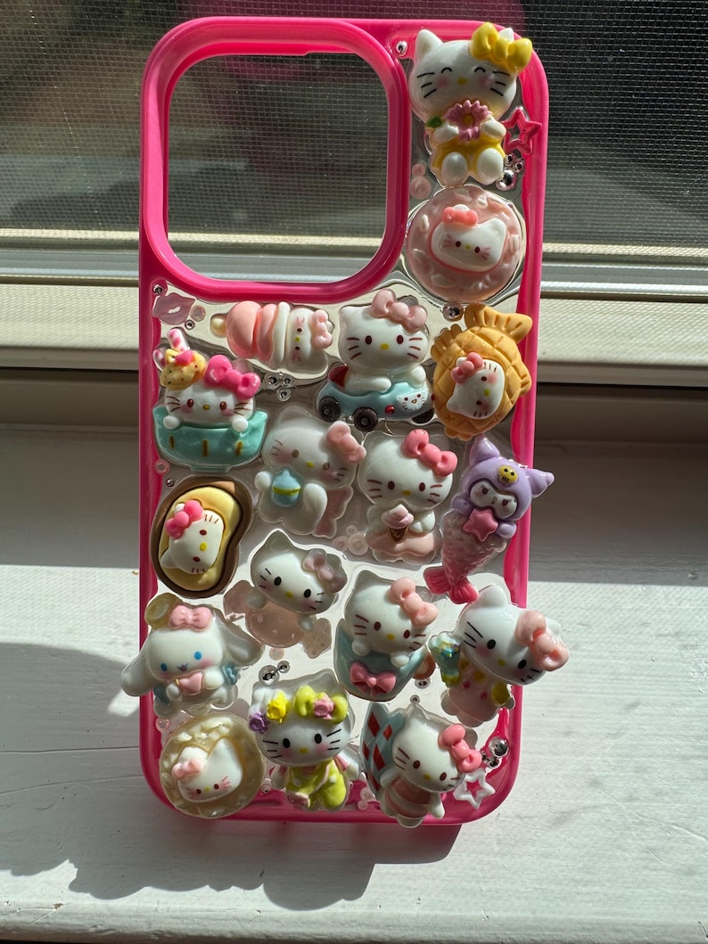 May include: A pink phone case adorned with multiple 3D Hello Kitty figurines in diverse poses and outfits. The case has a clear background with the Hello Kitty figures attached, and a pink border. The figurines include bows, flowers, and other accessories.