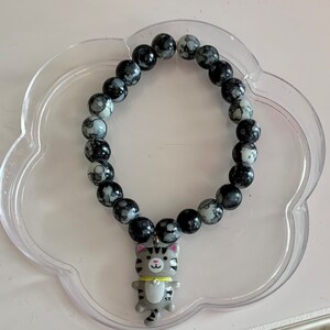 May include: A black and white beaded bracelet with a gray cat charm. The beads are round and have a marbled pattern.