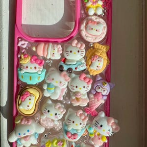 May include: A pink phone case embellished with multiple Hello Kitty charms. The charms feature various Hello Kitty figures, bows, and other decorative elements. The case is decorated with small, shiny embellishments, creating a playful and cute aesthetic.