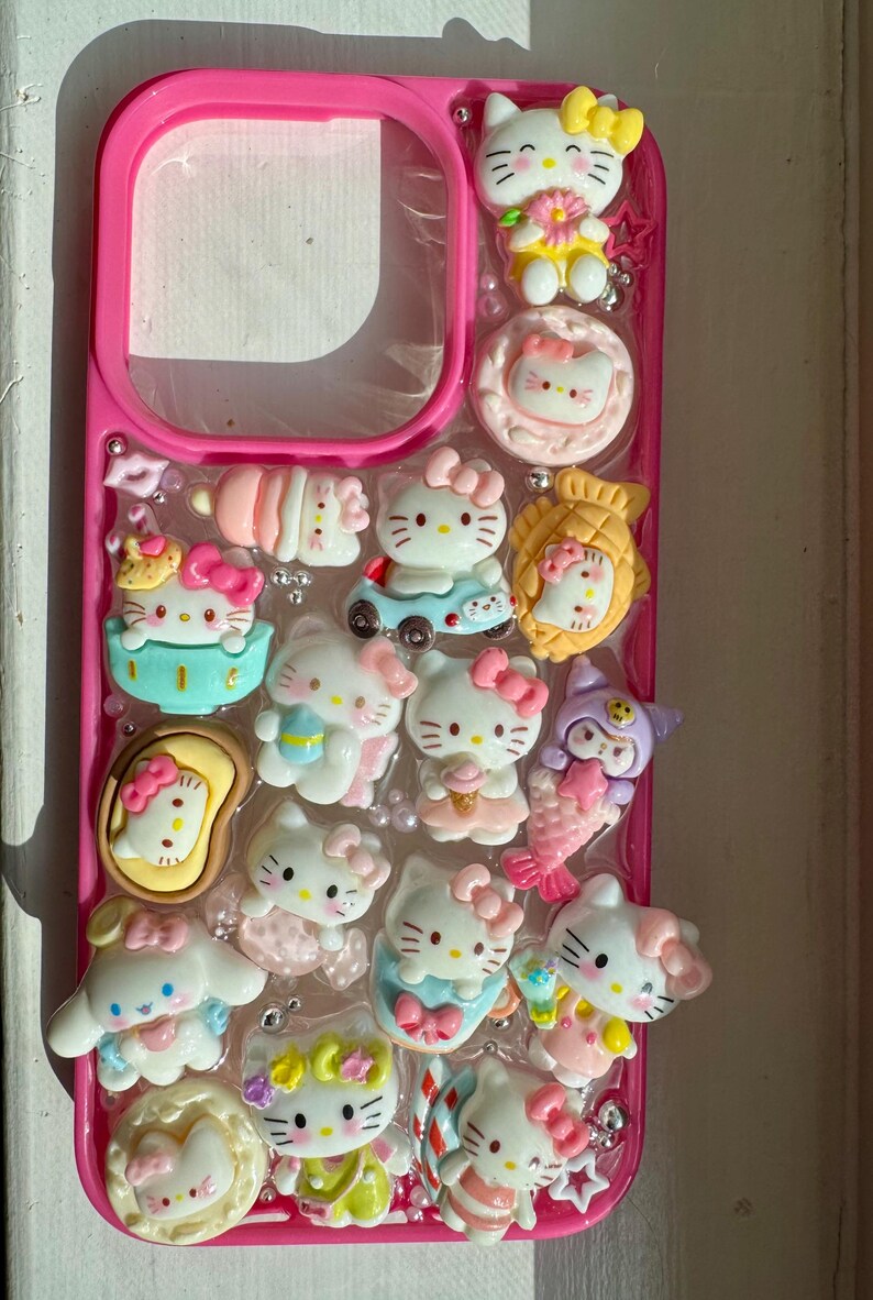 May include: A pink phone case embellished with multiple Hello Kitty charms. The charms feature various Hello Kitty figures, bows, and other decorative elements. The case is decorated with small, shiny embellishments, creating a playful and cute aesthetic.