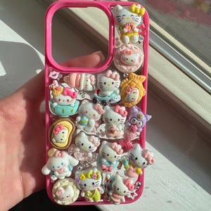 May include: A pink phone case decorated with multiple Hello Kitty character charms. The charms feature different poses and outfits, with pastel colours and glitter. The case is held in a hand near a window.